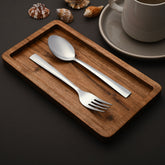 Stainless steel fork and spoon neatly arranged on wood tray beside seashells and ceramic cup.